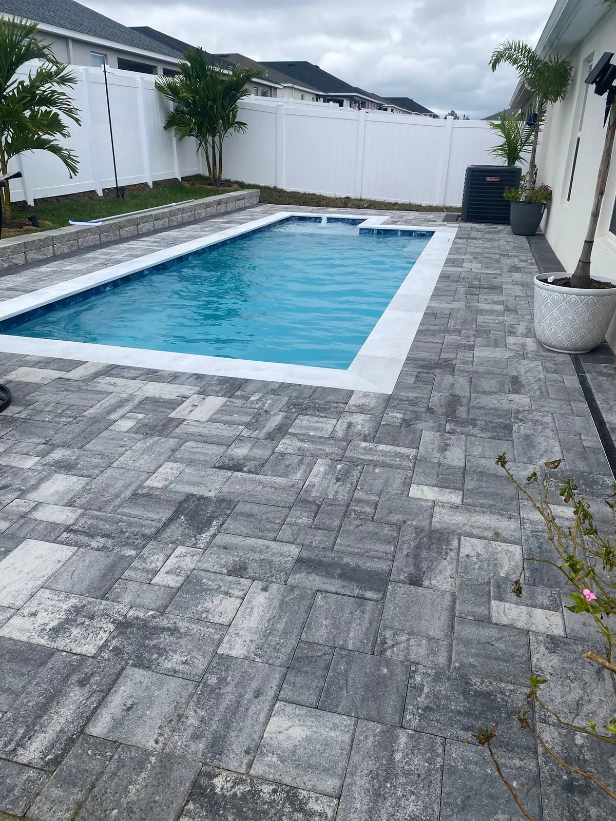Gray pavers surrounding a rectangular swimming pool, with white fencing in the background.